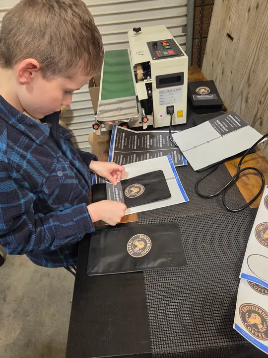 A child placing a Brotherhood Coffee label on a black coffee bag