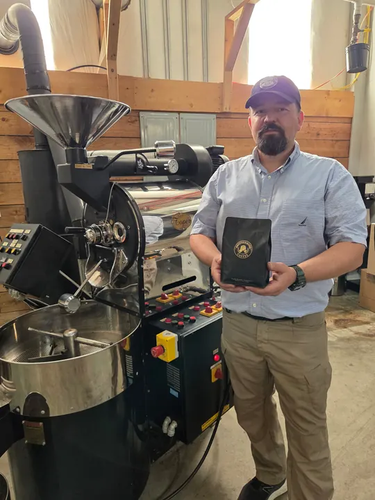 Brotherhood Coffee roaster holding a black coffee bag beside the roaster