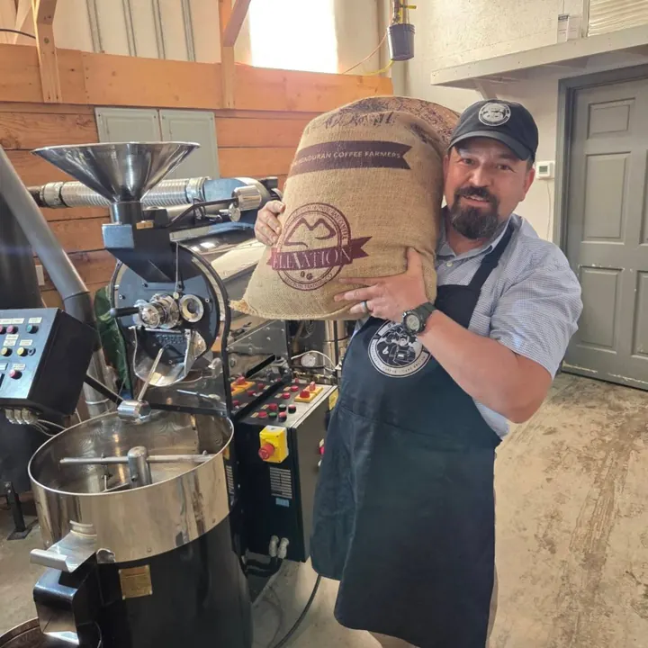 Brotherhood Coffee roaster holding a burlap coffee sack beside the roaster